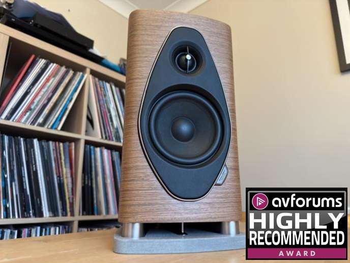 Wooden veneer bookshelf speaker on a metal stand in a living room, with vinyl records on shelves behind and an Avforums Highly Recommended Award badge in the corner.